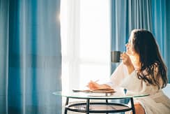 woman drinks coffee and sitting at glass table in spacious bedroom in sunny lazy morning.