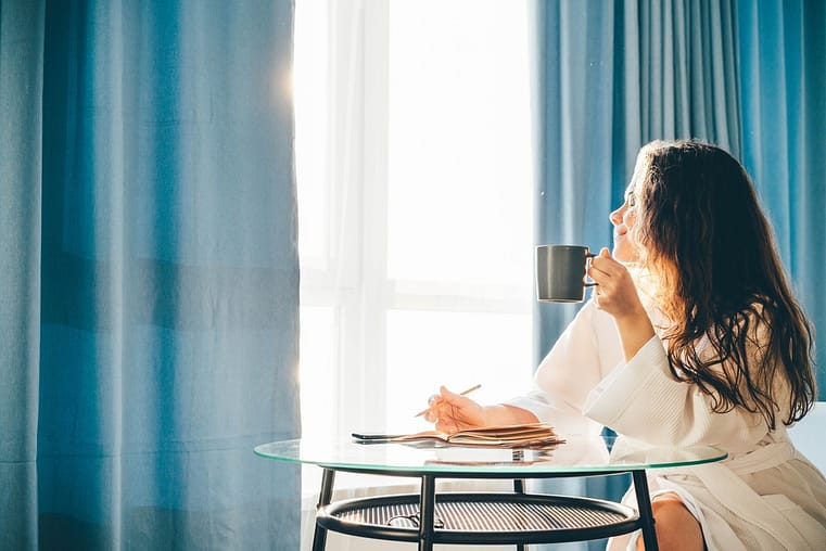 woman drinks coffee and sitting at glass table in spacious bedroom in sunny lazy morning.